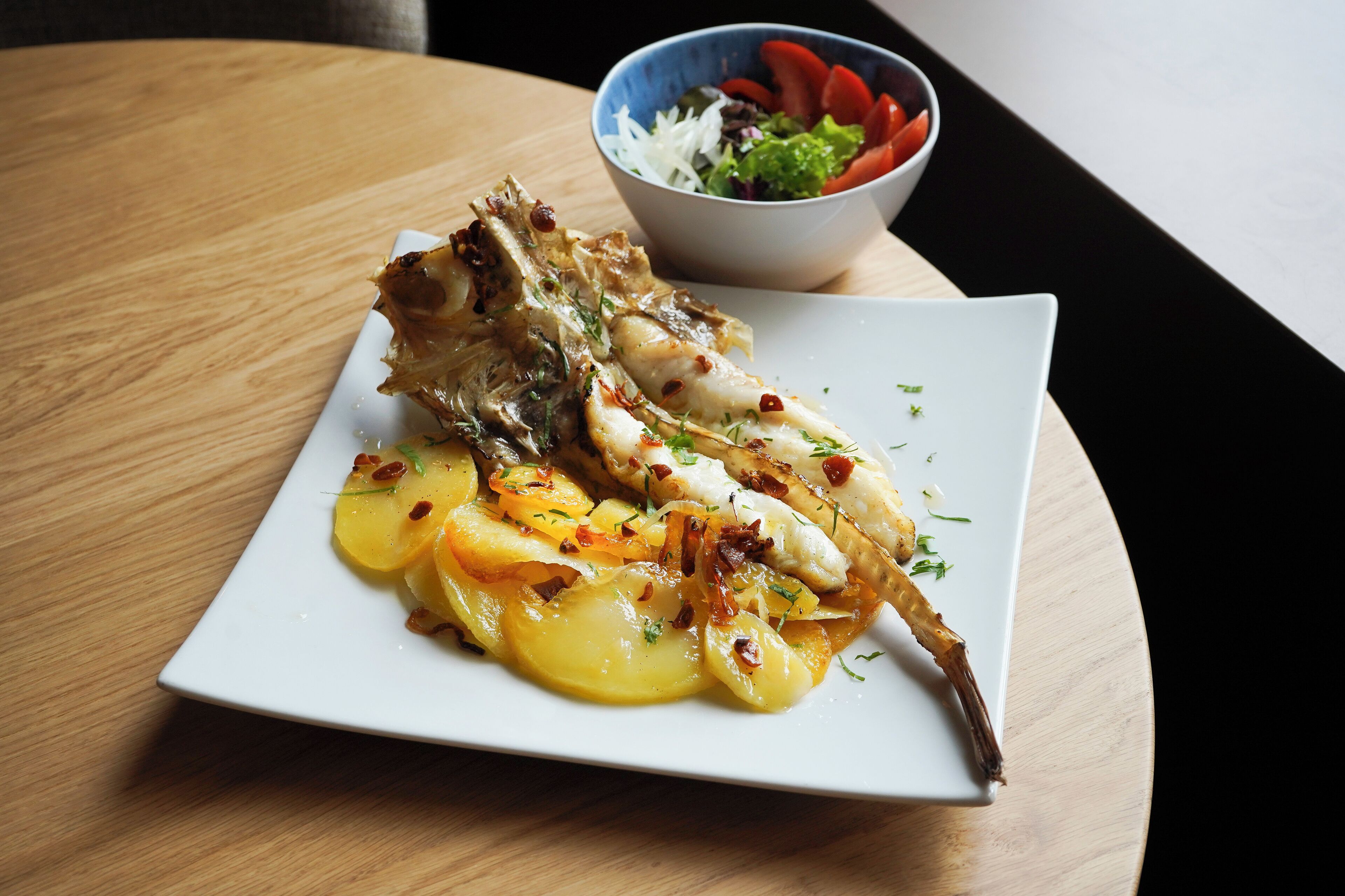 A plate of food with a fish and potatoes, and a bowl of salad on a wooden table.