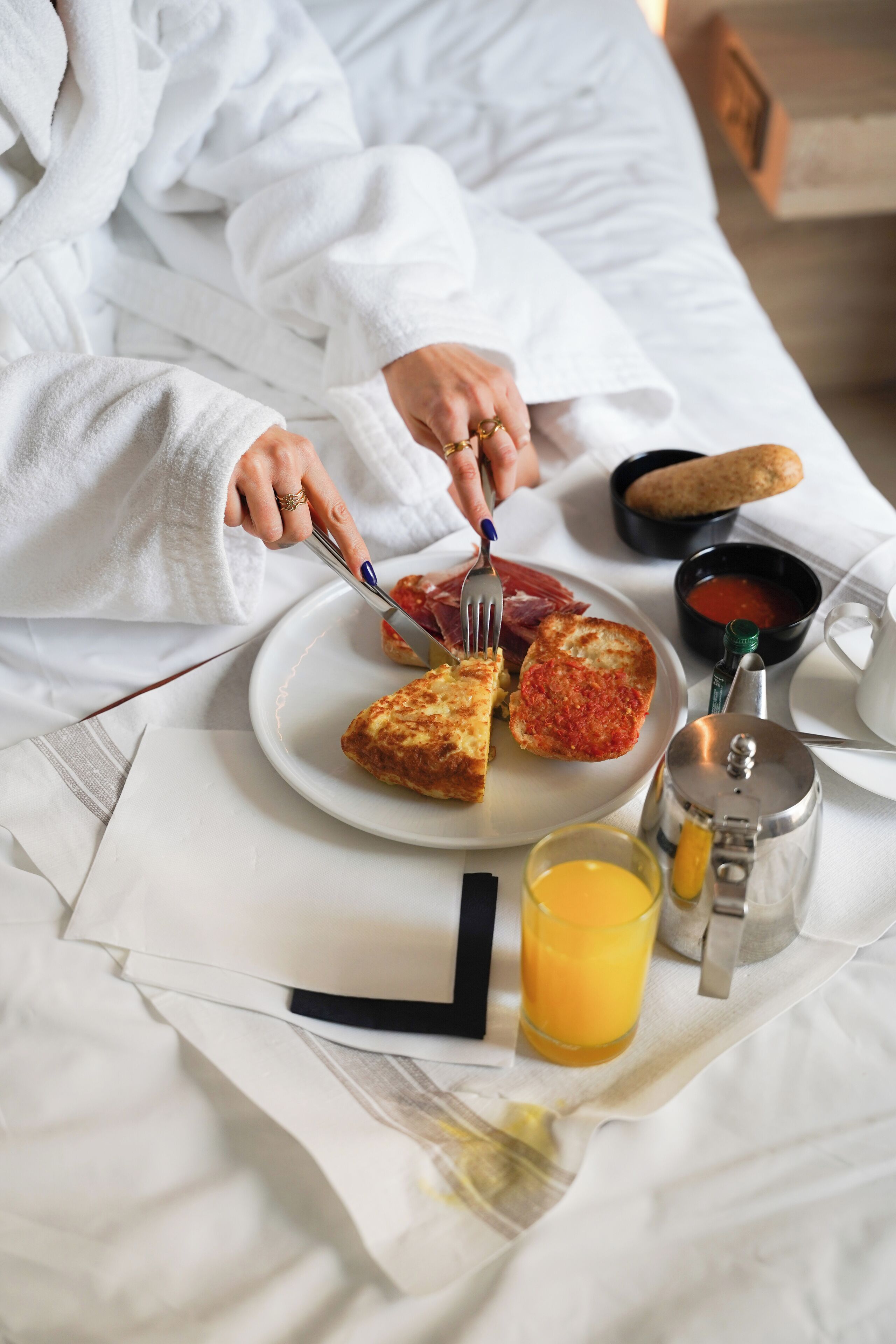 A person in a white robe sitting on a bed with a breakfast tray containing toast, a slice of pizza, a bowl of soup, a glass of orange juice, and a coffee pot.
