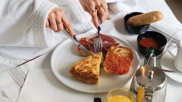 A person in a white robe sitting on a bed with a breakfast tray containing toast, a slice of pizza, a bowl of soup, a glass of orange juice, and a coffee pot.
