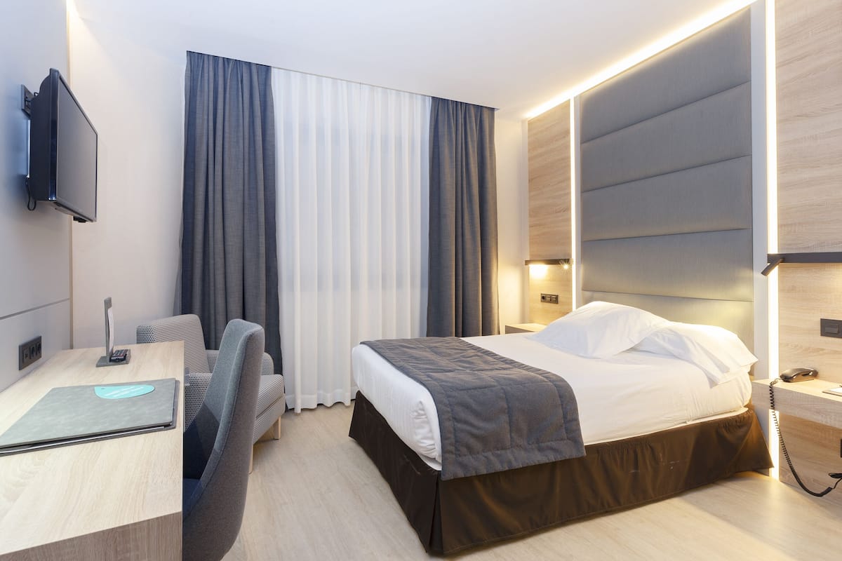 Moderni hotellihuone, jossa on sänky, yöpöytä, lamppu ja työpöytä, jossa on kannettava tietokone.