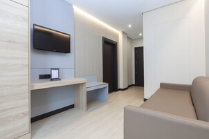Una moderna camera d'albergo con televisione a schermo piatto fissata al muro, una panca in legno, un divano e una porta che conduce in un'altra stanza.