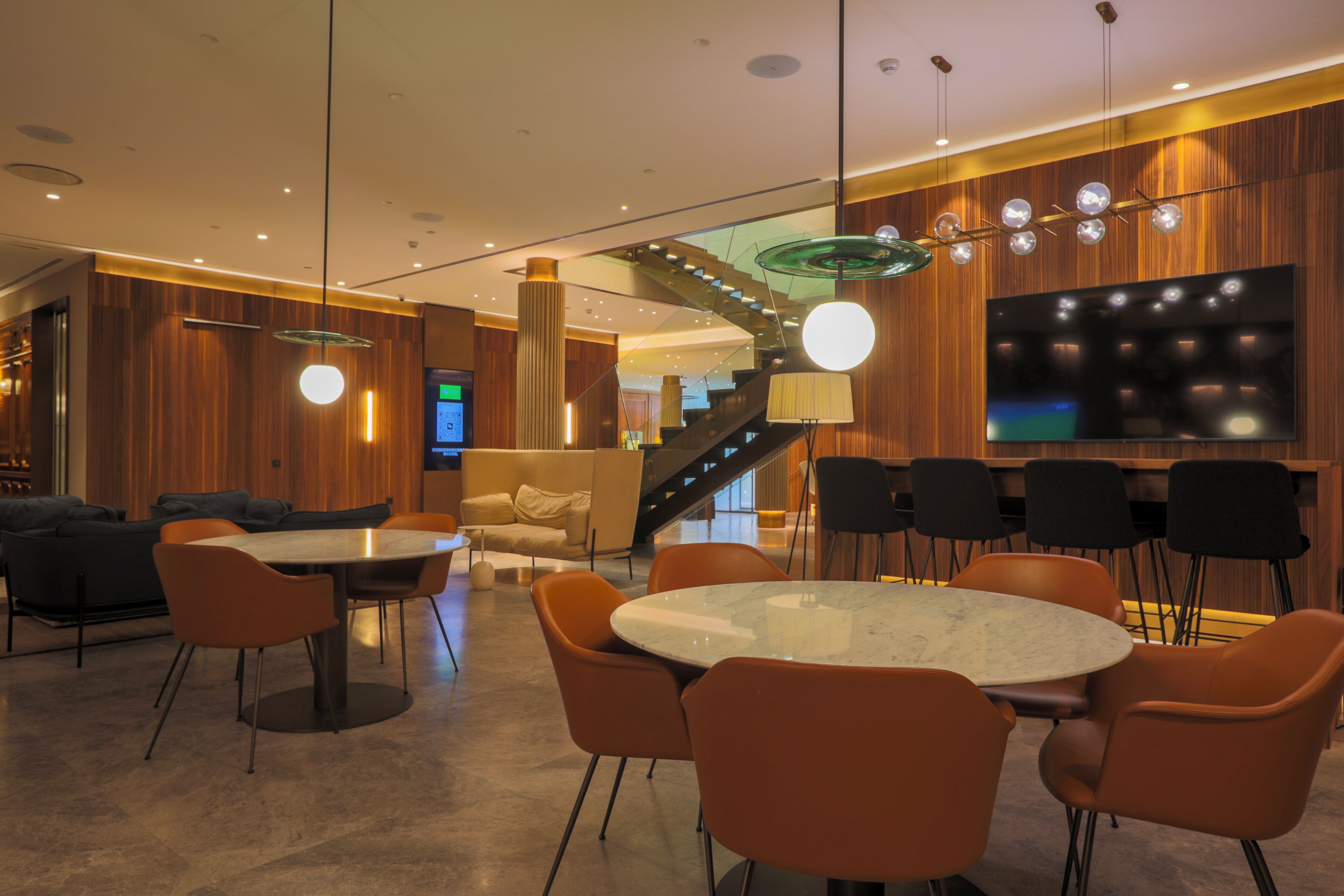 A modern hotel lobby with a round marble table, orange chairs, a staircase, and a large flat-screen TV.