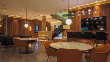 Eine moderne Hotelhalle mit einem runden Marmortisch, orangefarbenen Stühlen, einer Treppe und einem großen Flachbildfernseher.