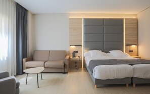 A modern hotel room with a bed, a sofa, a small table, and a nightstand with a lamp.