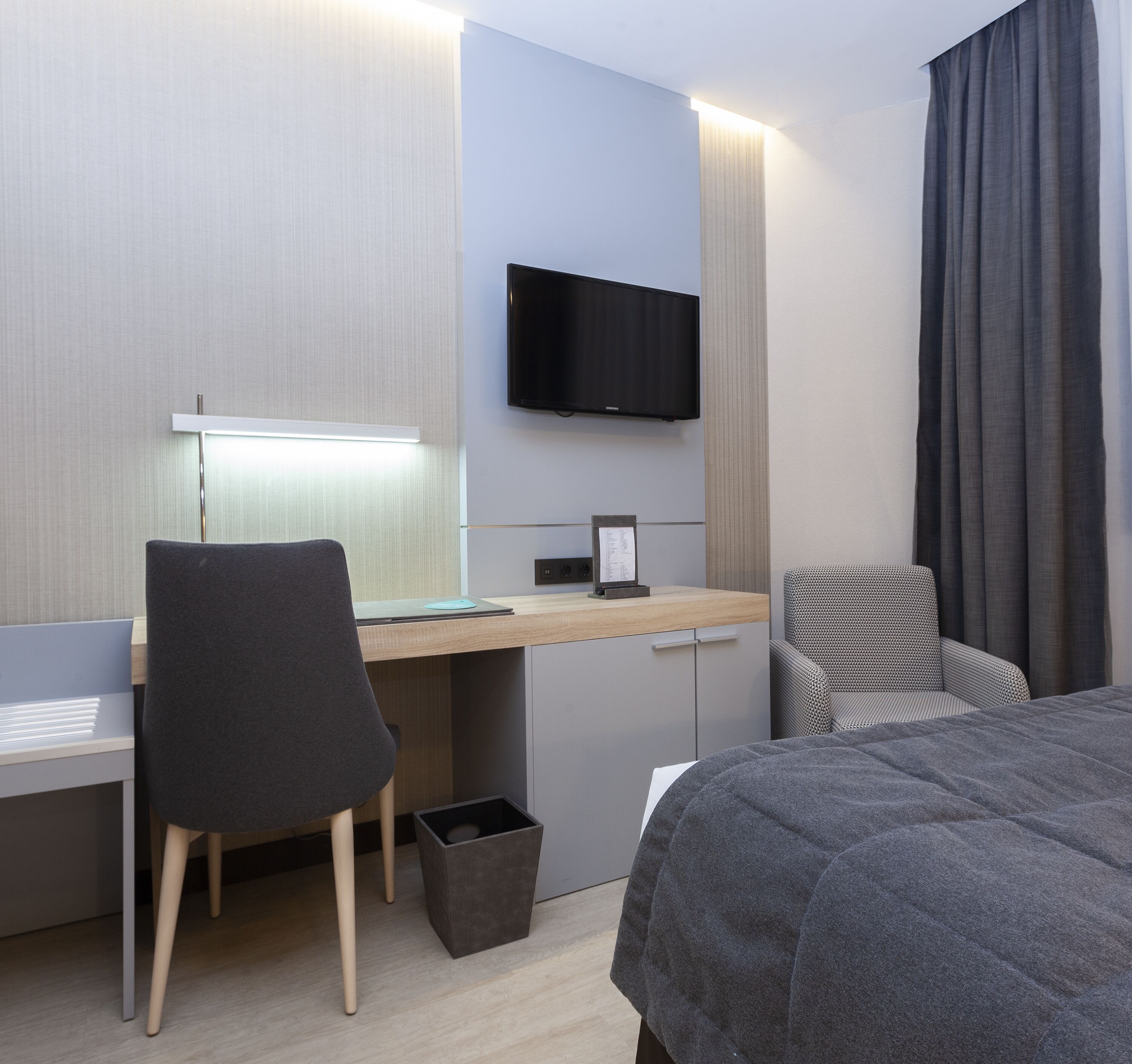 A modern hotel room with a desk, chair, bed, and a flat-screen TV mounted on the wall.