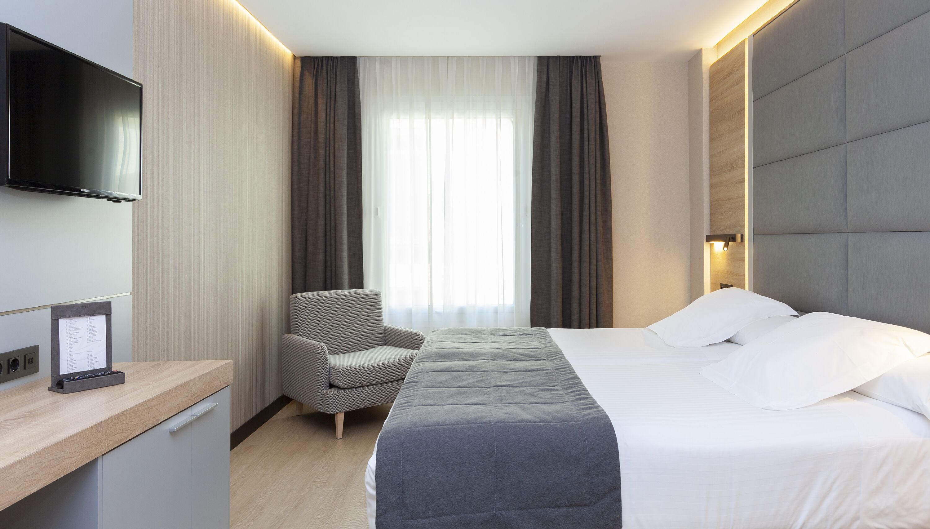 Habitación de hotel con cama, una silla, escritorio con televisor de pantalla plana y ventana con cortinas.