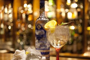 Eine Bar mit einem Glas, das einen Cocktail, Eiswürfel und Garnituren enthält, und einer mit 'GIN' beschrifteten Flasche im Hintergrund.
