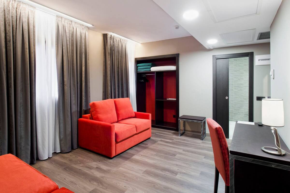 A modern hotel room with a red sofa, a small table with a lamp, a wardrobe, and a door with a mirror.
