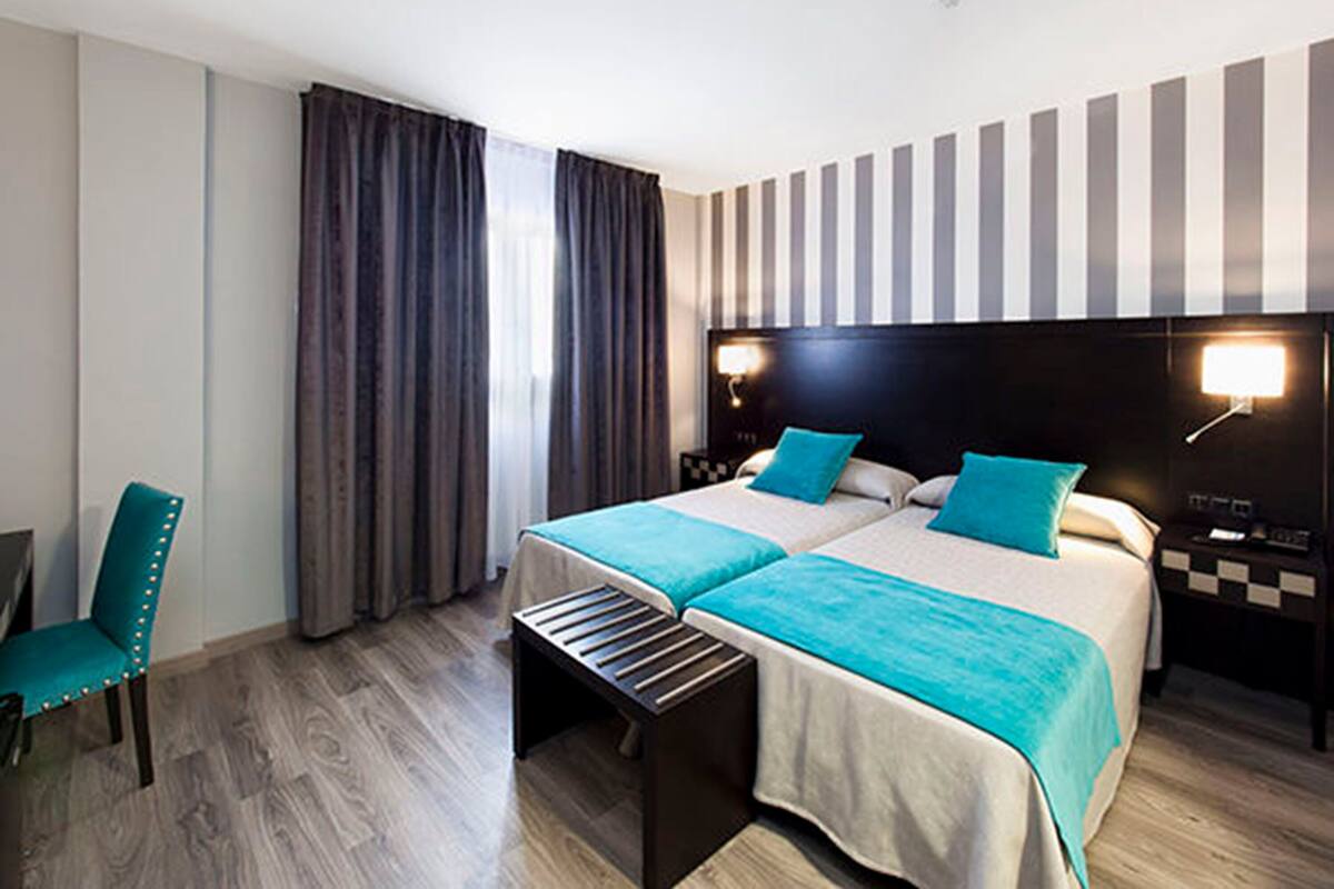 A hotel room with two beds, a bench, a chair, and a striped wall.