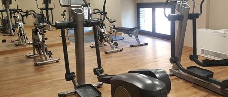 A gym with various exercise machines, including treadmills and elliptical trainers, arranged on a wooden floor.
