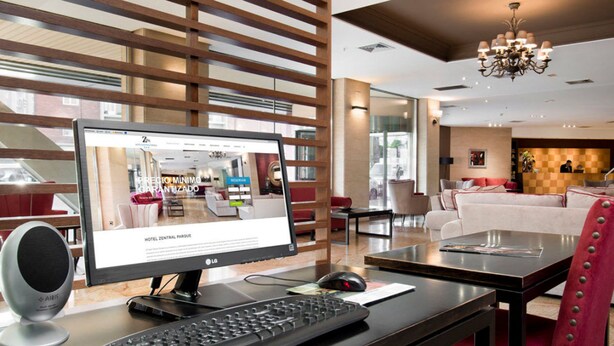 A modern hotel lobby with a computer monitor displaying a hotel website, a desk with a keyboard and mouse, a red upholstered chair, and a chandelier. - Hotel Zentral Parque (Valladolid)