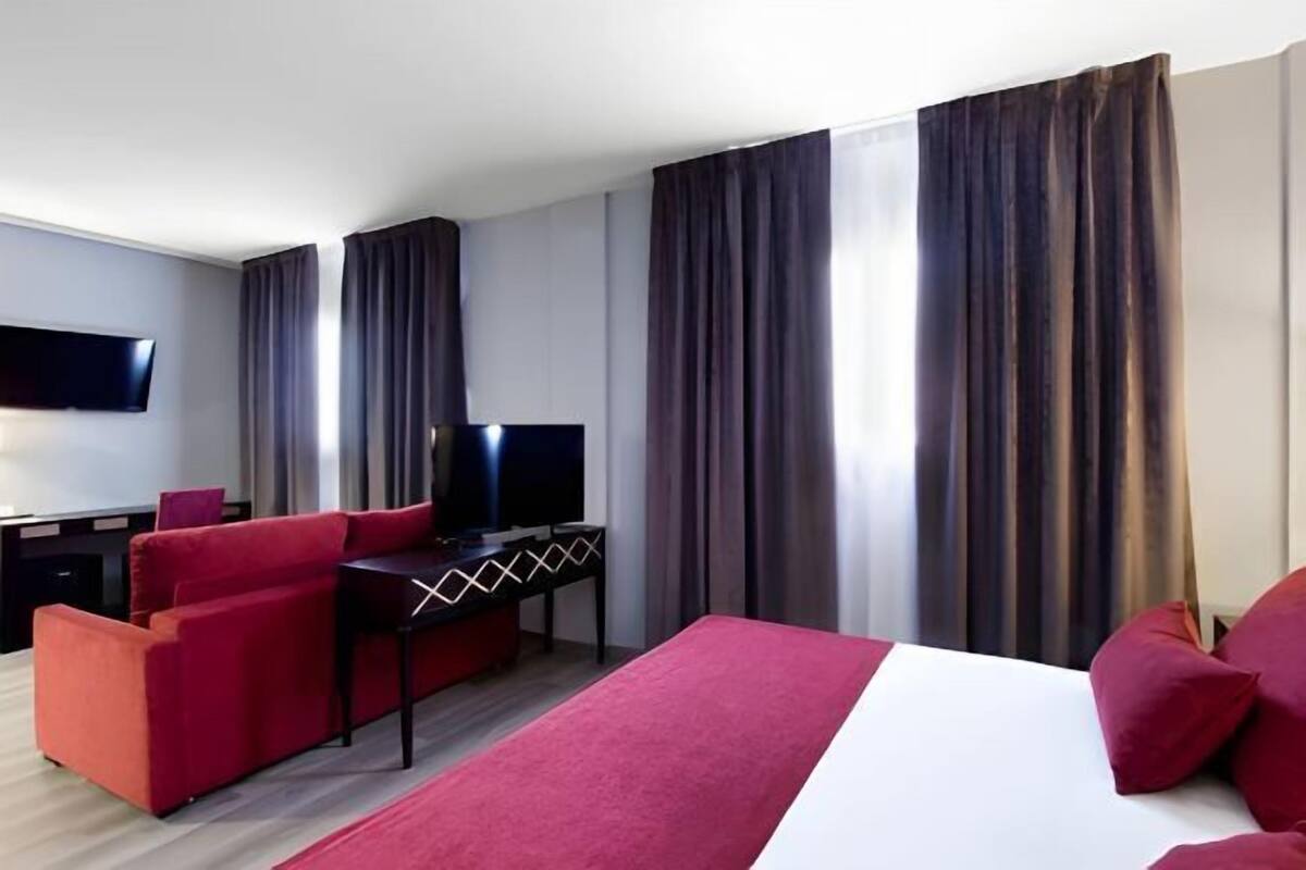 A hotel room with a bed, a red sofa, a desk with a flat-screen TV, and large curtains.