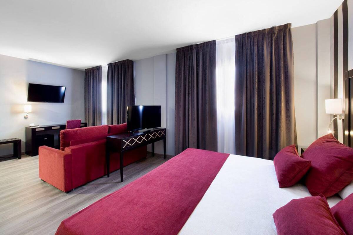 A hotel room with a large bed, a red sofa, a desk with a TV, and curtains.