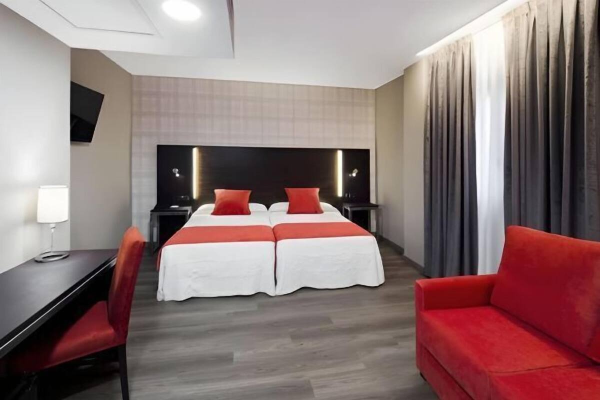 A hotel room with a bed, two bedside tables, a desk, a chair, a red sofa, and a TV mounted on the wall.