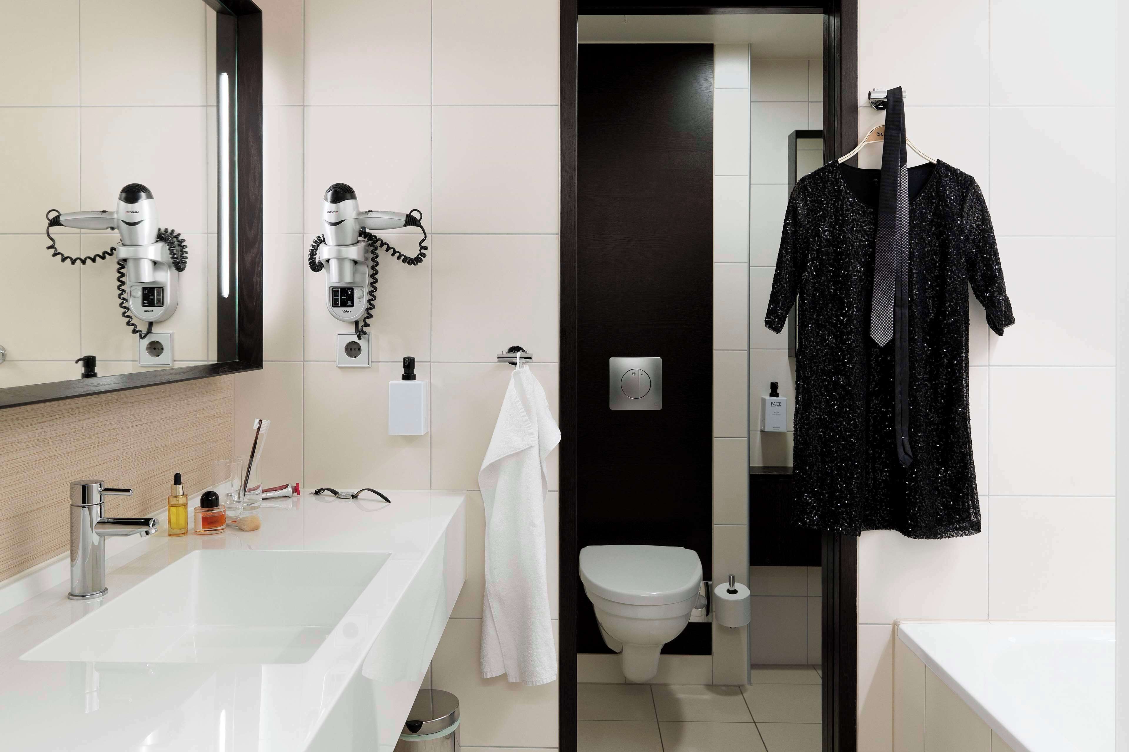 A modern bathroom with a large white sink, two hair dryers mounted on the wall, a toilet, and a black robe hanging on a rack.