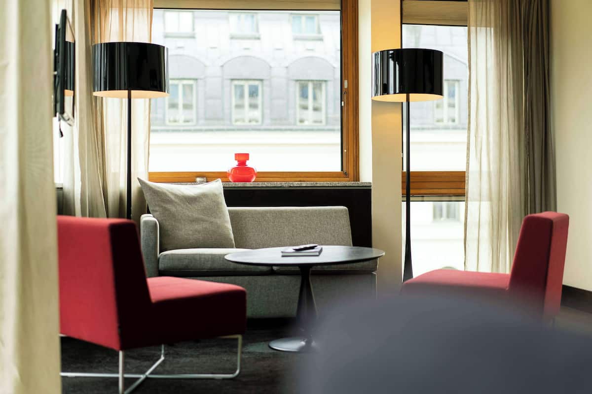 Ein modernes Hotelzimmer mit grauem Sofa, roten Sesseln, einem kleinen runden Tisch und zwei schwarzen Stehlampen.