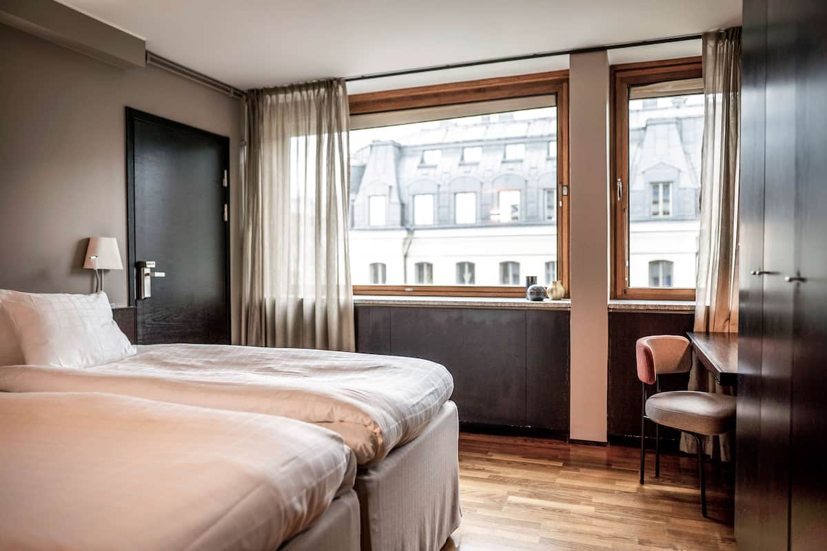 Ein Hotelzimmer mit einem großen Bett, einem Schreibtisch und Blick auf Gebäude durch ein Fenster.