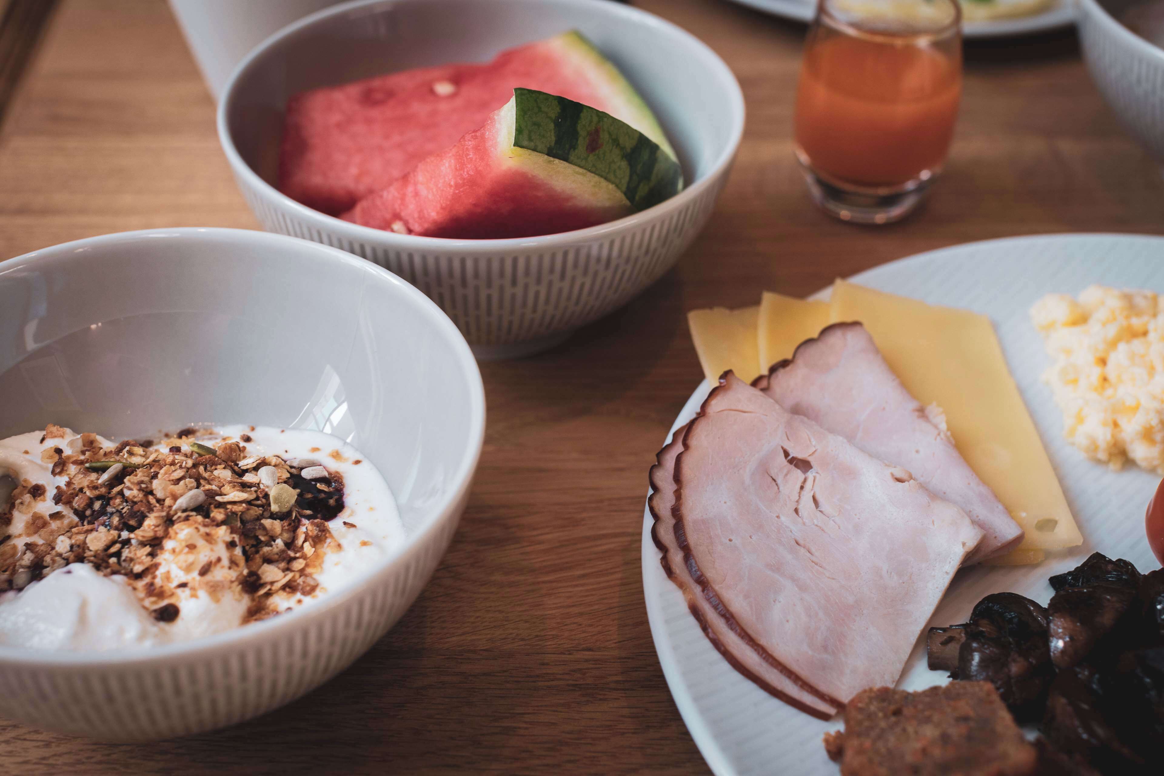 Un desayuno con tazones de yogur y granola, un plato con jamón, queso y champiñones, y un vaso de zumo.