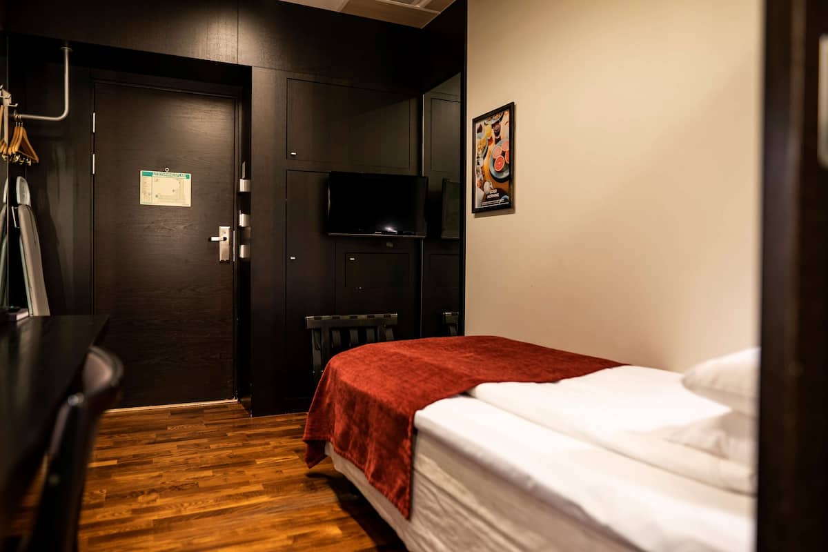 Ein Hotelzimmer mit Holzboden, einem Bett mit weißer Matratze und roter Decke, einer dunklen Tür mit einem Schild, einem an der Wand montierten Flachbildfernseher und einem Heizkörper.