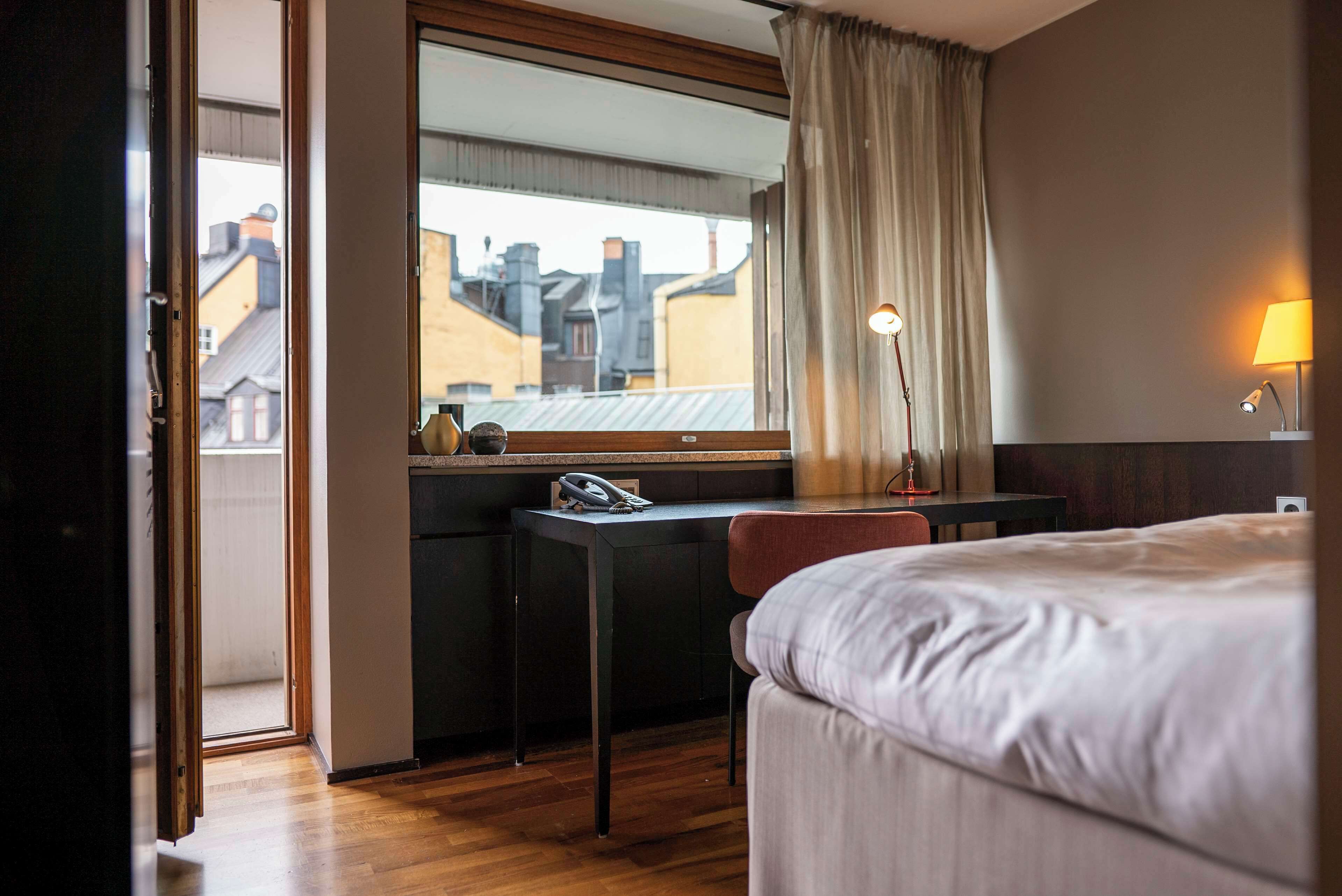 Ett hotellrum med en säng, ett skrivbord, en stol och utsikt över byggnader genom ett fönster.