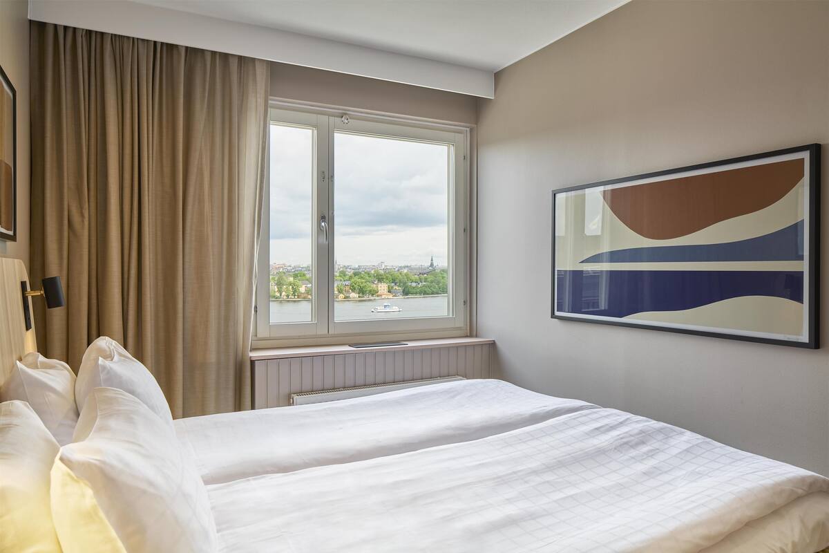 A hotel room with a large bed, a window with a view of a cityscape, and a painting on the wall.