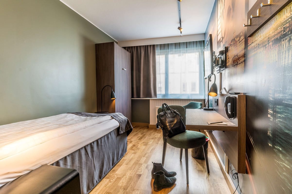 A compact hotel room with a single bed, a desk with a chair, a small table, a lamp, and a large window.