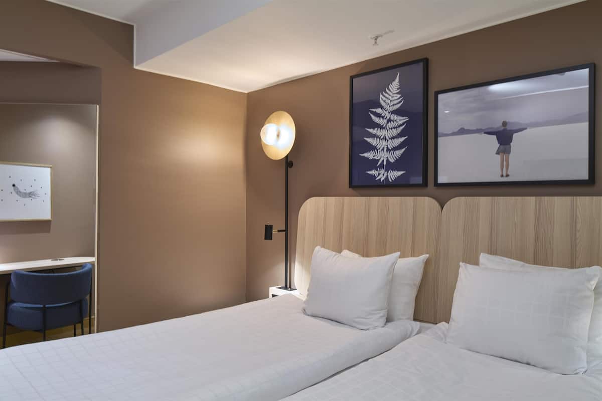 A hotel room with a bed, a desk, a chair, a lamp, and two framed pictures on the wall.