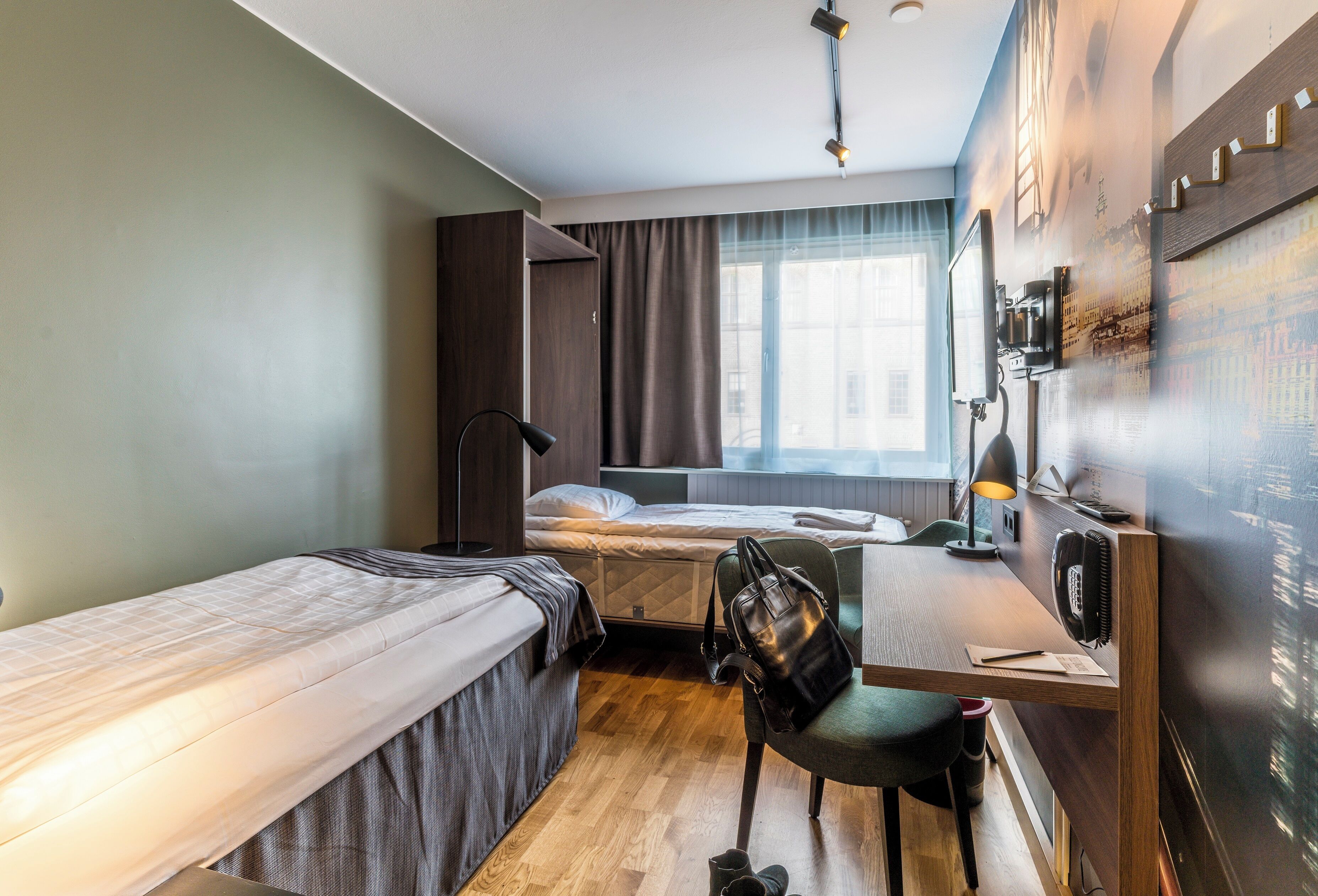 A compact hotel room with two single beds, a desk with a chair, a desk lamp, and a large window with curtains.