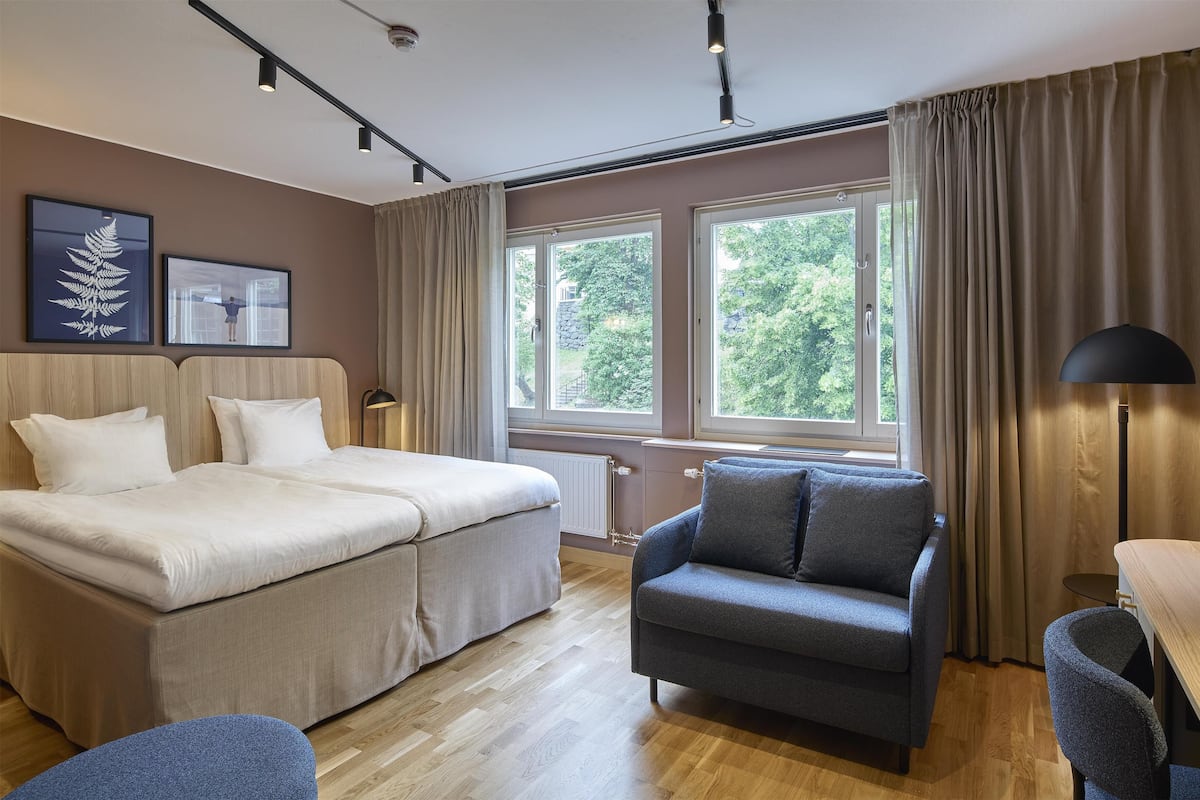 A hotel room with a bed, a chair, a desk, and large windows with curtains.