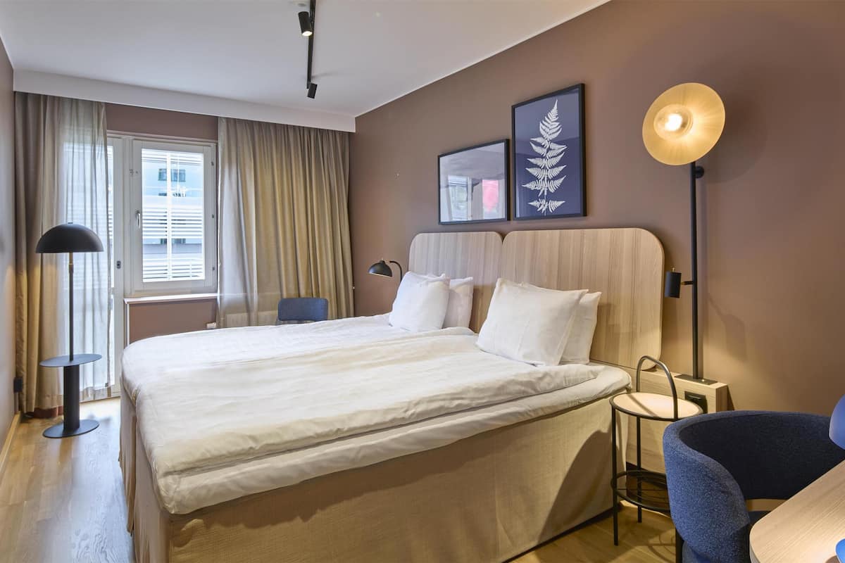 A hotel room with a large bed, a desk, a chair, a lamp, and a window with curtains.