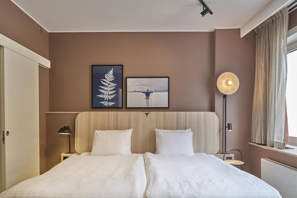 A hotel room with a large bed, two framed pictures on the wall, a bedside lamp, and a window with curtains.