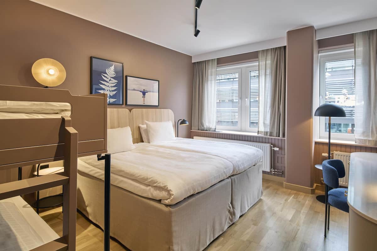 A hotel room with a bed, a desk, a chair, a window with curtains, and a radiator.