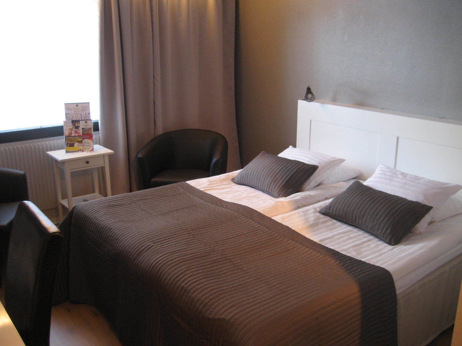 A hotel room with a bed, a chair, a small table with a magazine, and a window with curtains.