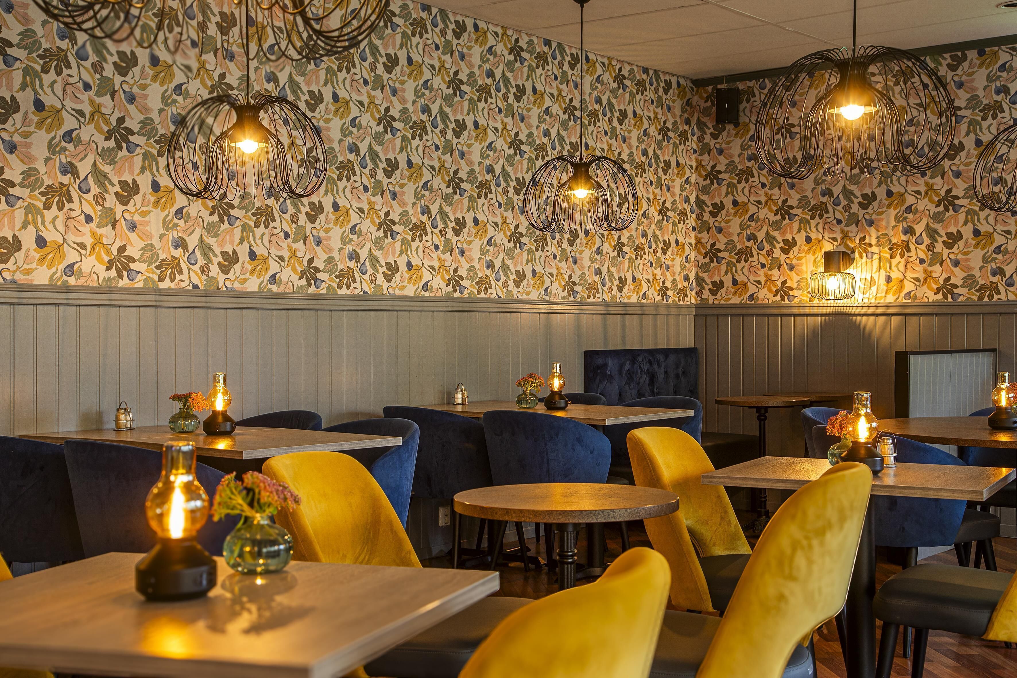 Ein Restaurantinnenraum mit strukturiertem Wandpapier, Pendelleuchten und Tischen mit Stühlen.