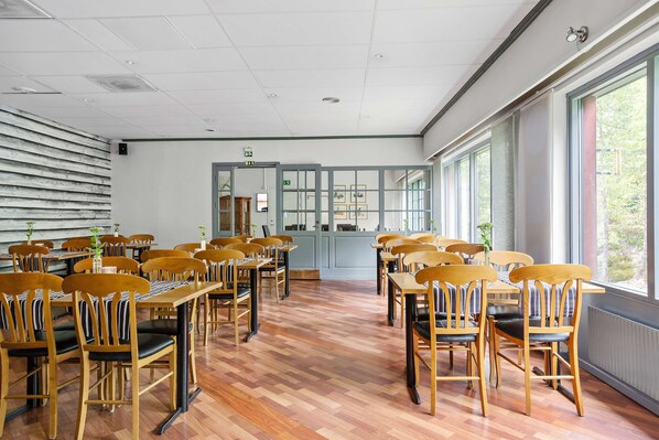 A spacious room with wooden chairs and tables arranged for dining, large windows providing natural light, and a view of greenery outside.