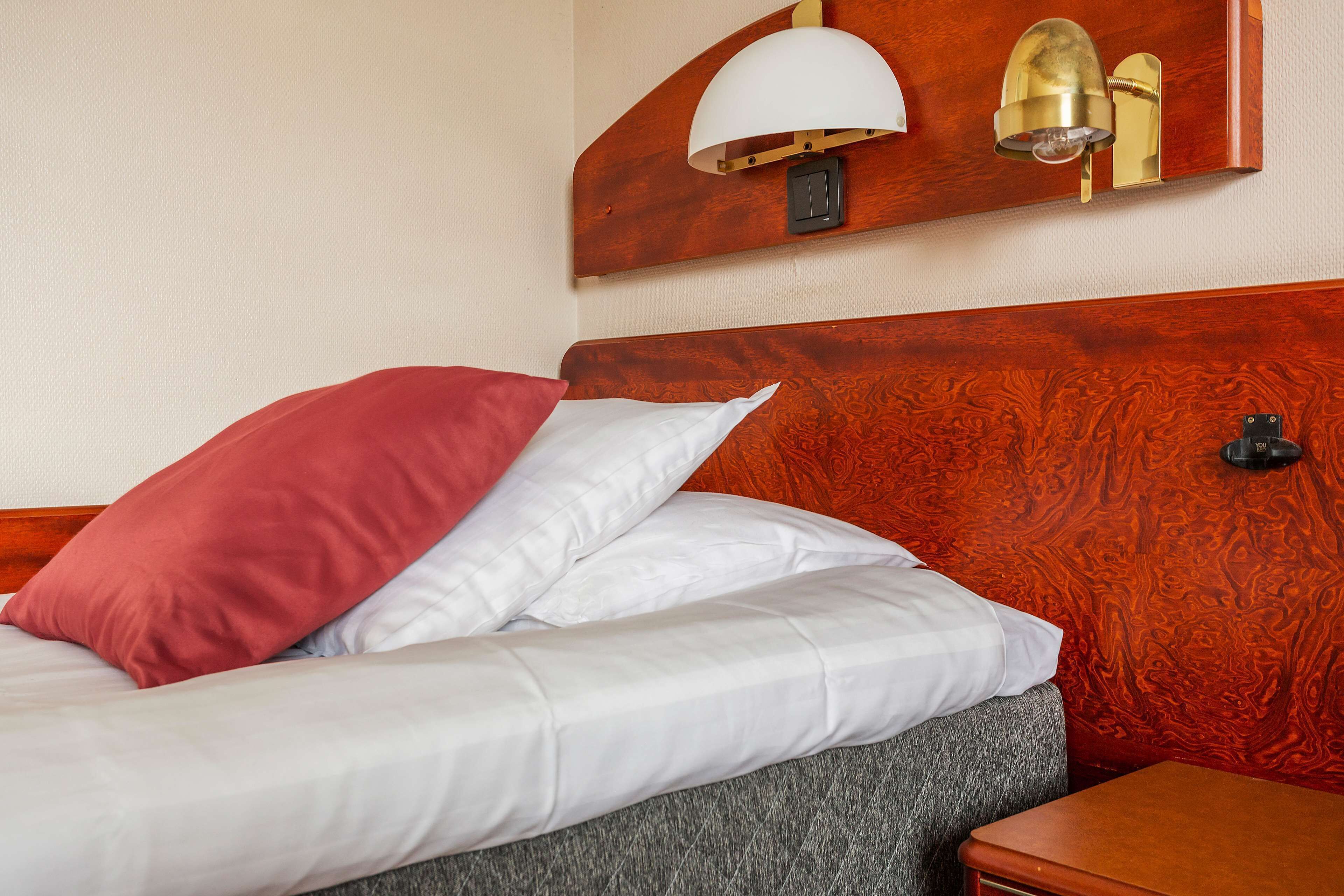 A neatly made bed with a red and white pillow, a wooden headboard, and a wall-mounted lamp.