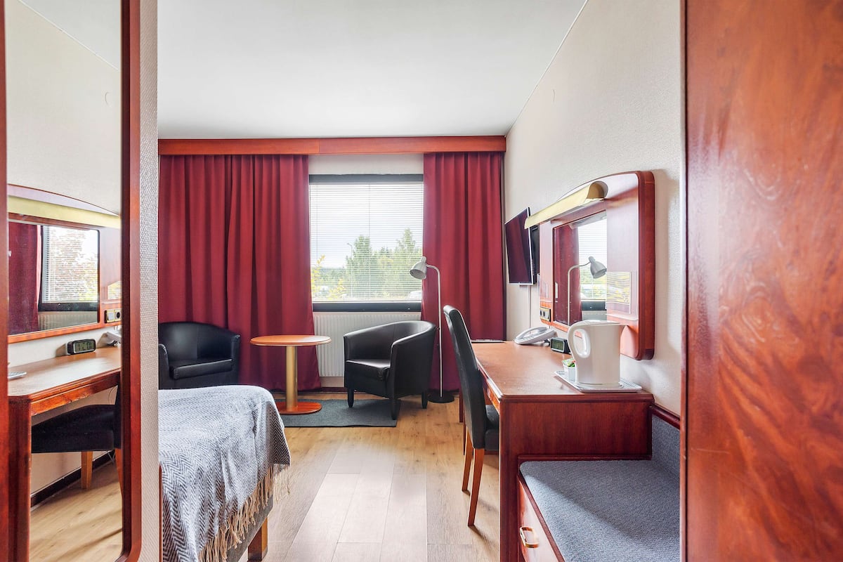 A hotel room with a wooden desk, two chairs, a small table, a TV, and a window with curtains.