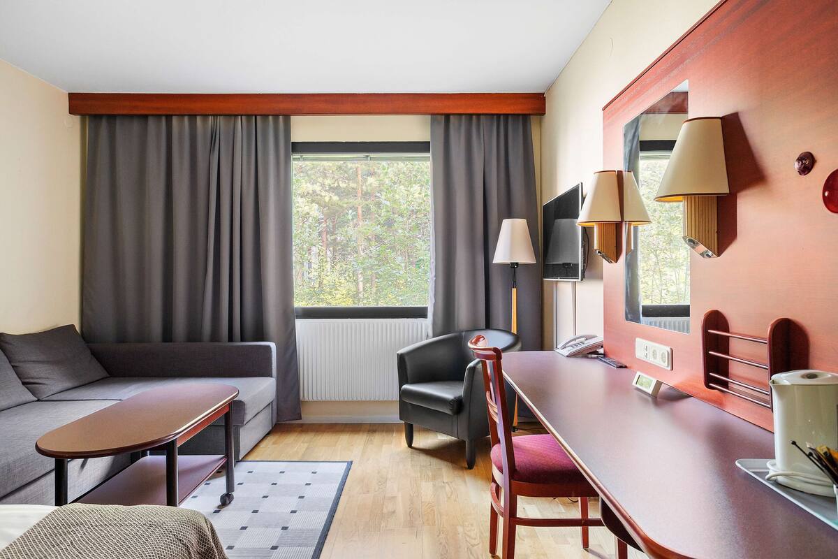 A modern hotel room with a sofa, a small table, a chair, a desk, a lamp, a mirror, and a view of trees through the window.