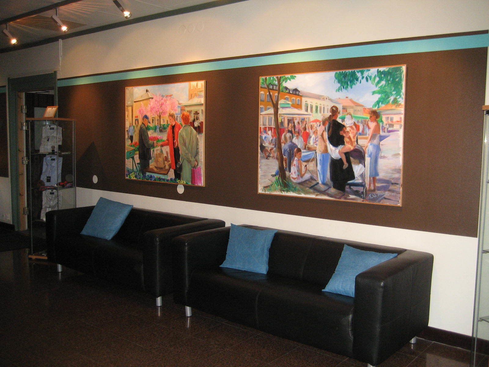 A lobby with two black leather sofas with blue cushions, a glass display case with folded clothes, and two large paintings depicting outdoor scenes.