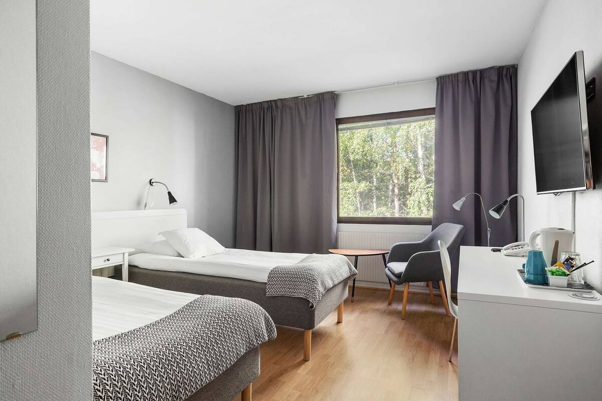 A hotel room with two beds, a desk, a chair, a TV, and a window with curtains.