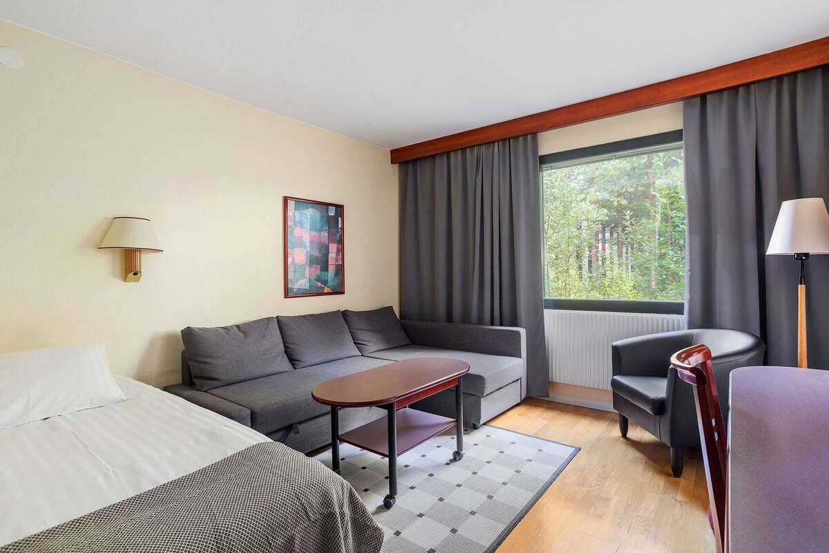 A hotel room with a bed, a sofa, a small table, a chair, a lamp, a painting, and a window with curtains.