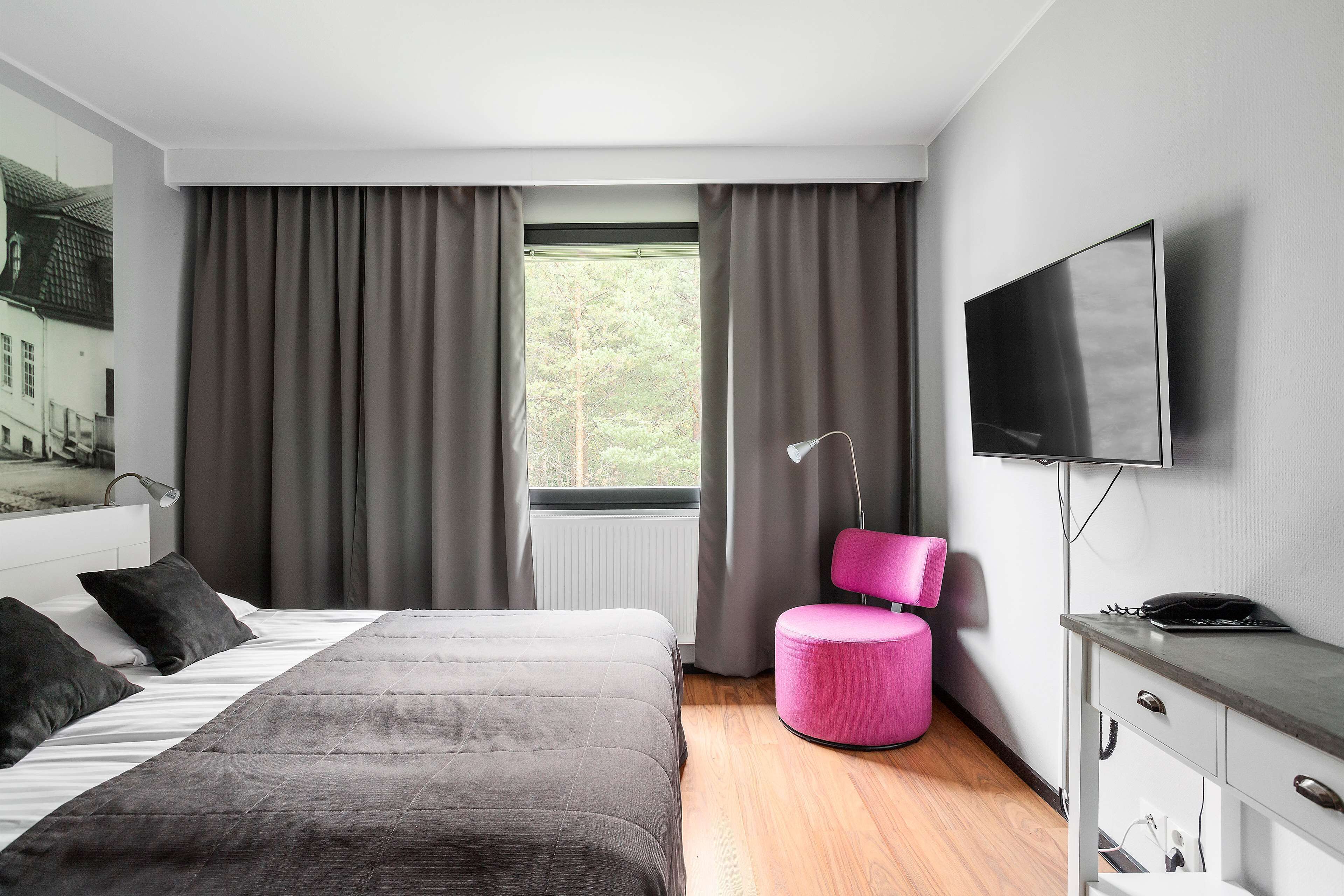 Moderni hotellihuone, jossa on suuri sänky, työpöytä, jossa on litteänäyttöinen televisio, vaaleanpunainen tuoli ja ikkuna, jossa on verhot.