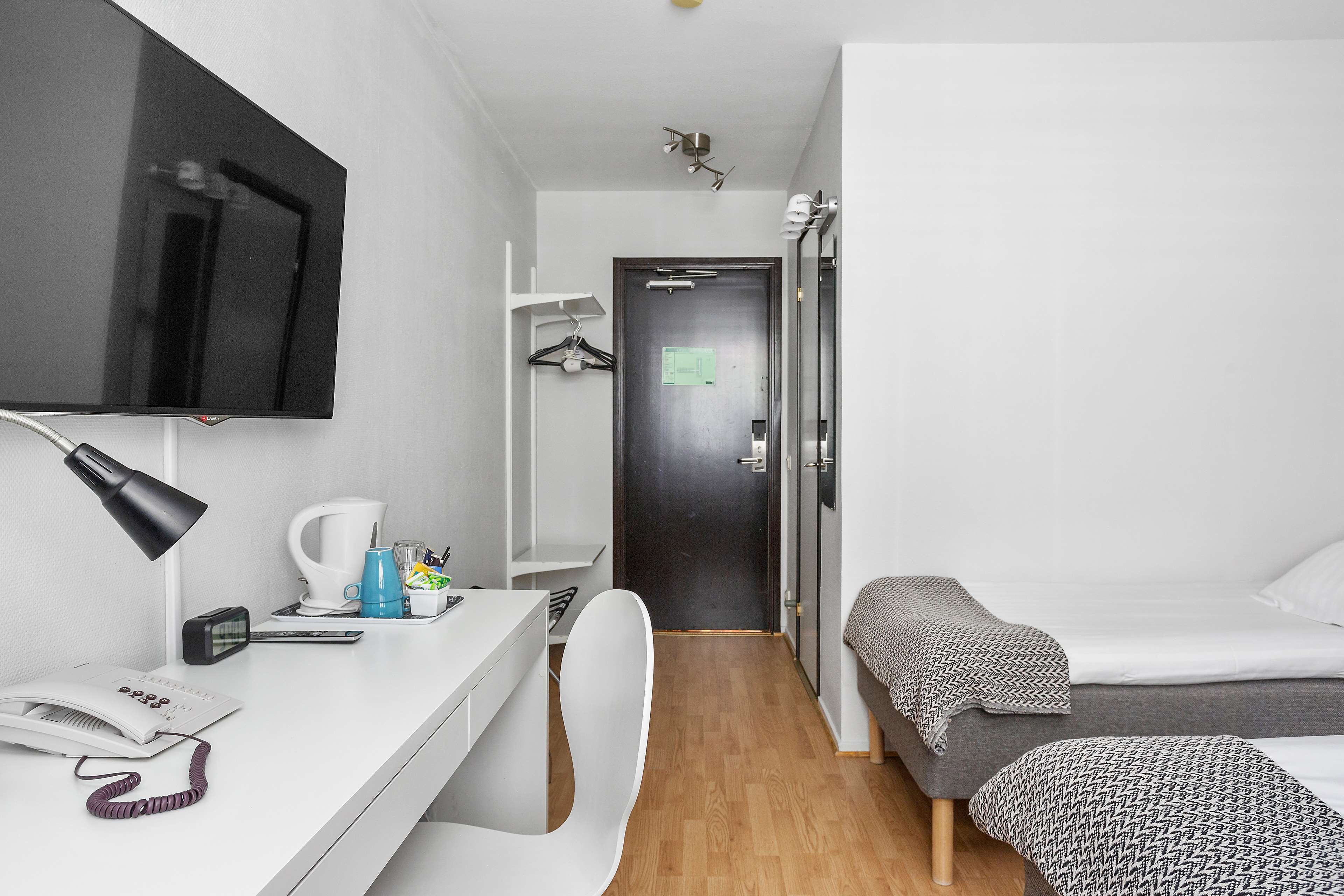 A compact hotel room with a single bed, a desk with a telephone, a chair, a TV mounted on the wall, and a small kitchenette area.
