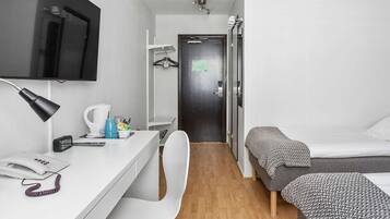 A compact hotel room with a single bed, a desk with a telephone, a chair, a TV mounted on the wall, and a small kitchenette area.