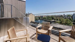 A rooftop terrace with wooden chairs and a table, overlooking a cityscape and a river. - Scandic Strömmen (Norrköping)