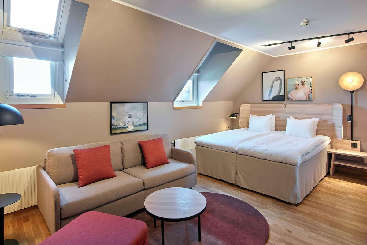 A modern bedroom with a bed, a sofa, a small table, and a red ottoman. There are two windows providing natural light and a ceiling light fixture.