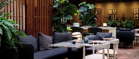 A modern lounge area with a sofa, chairs, tables, and large potted plants.