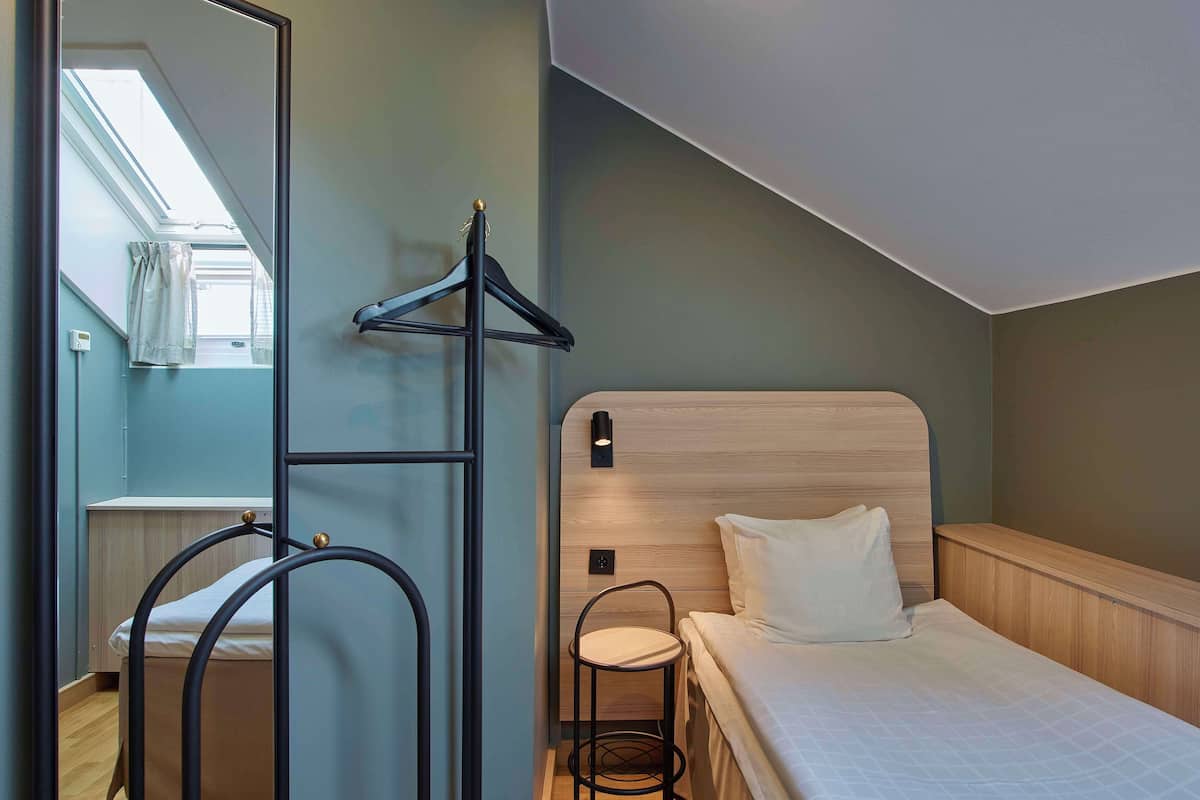 A compact bedroom with a bed, a small chair, a coat rack, and a mirror.
