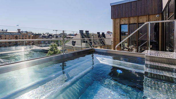 A rooftop swimming pool with clear water, surrounded by buildings and a wooden structure with large windows. - Scandic Strömmen (Norrköping)