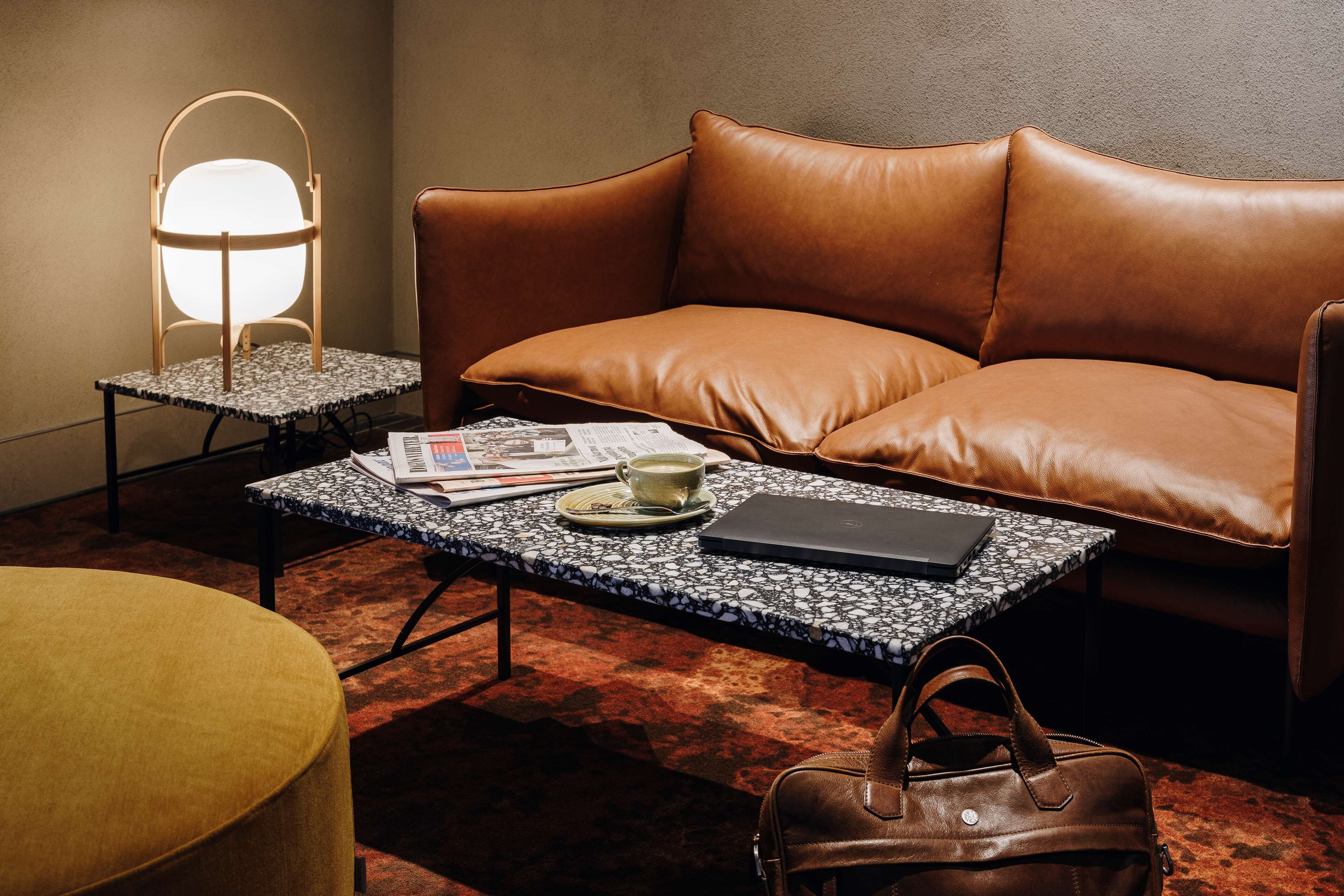 A cozy room with a leather sofa, a coffee table with a laptop and a cup, a lamp, and a handbag.